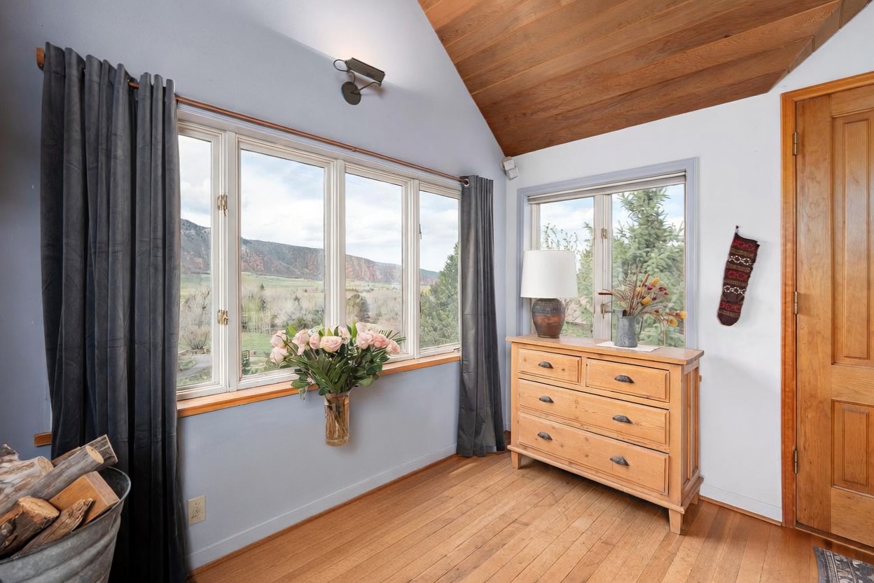 This bright sunroom features light blue walls, warm wood flooring, and a vaulted ceiling with exposed wood paneling. Large windows provide expansive views of the surrounding landscape, while a rustic wooden chest and a light wood dresser offer functional storage. The space is accented by a vase of fresh roses and a lamp, creating a cozy and inviting atmosphere.