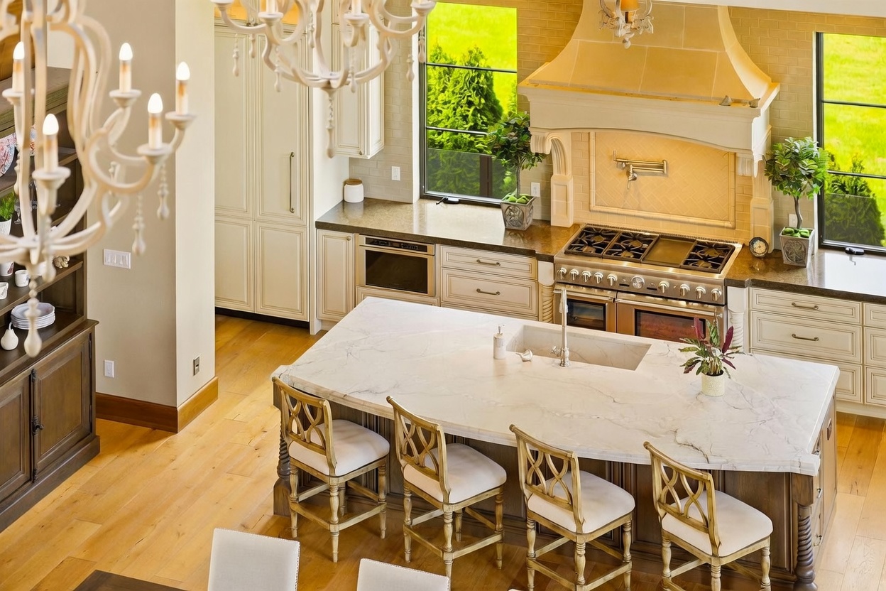 This high-angle shot captures a spacious, elegant kitchen featuring a large central island with a stone countertop and seating for four. The space is equipped with professional-grade stainless steel appliances, including a prominent range with a decorative hood, and is accented by light-toned cabinetry and warm wood flooring. Large windows provide ample natural light, creating an inviting and sophisticated atmosphere.