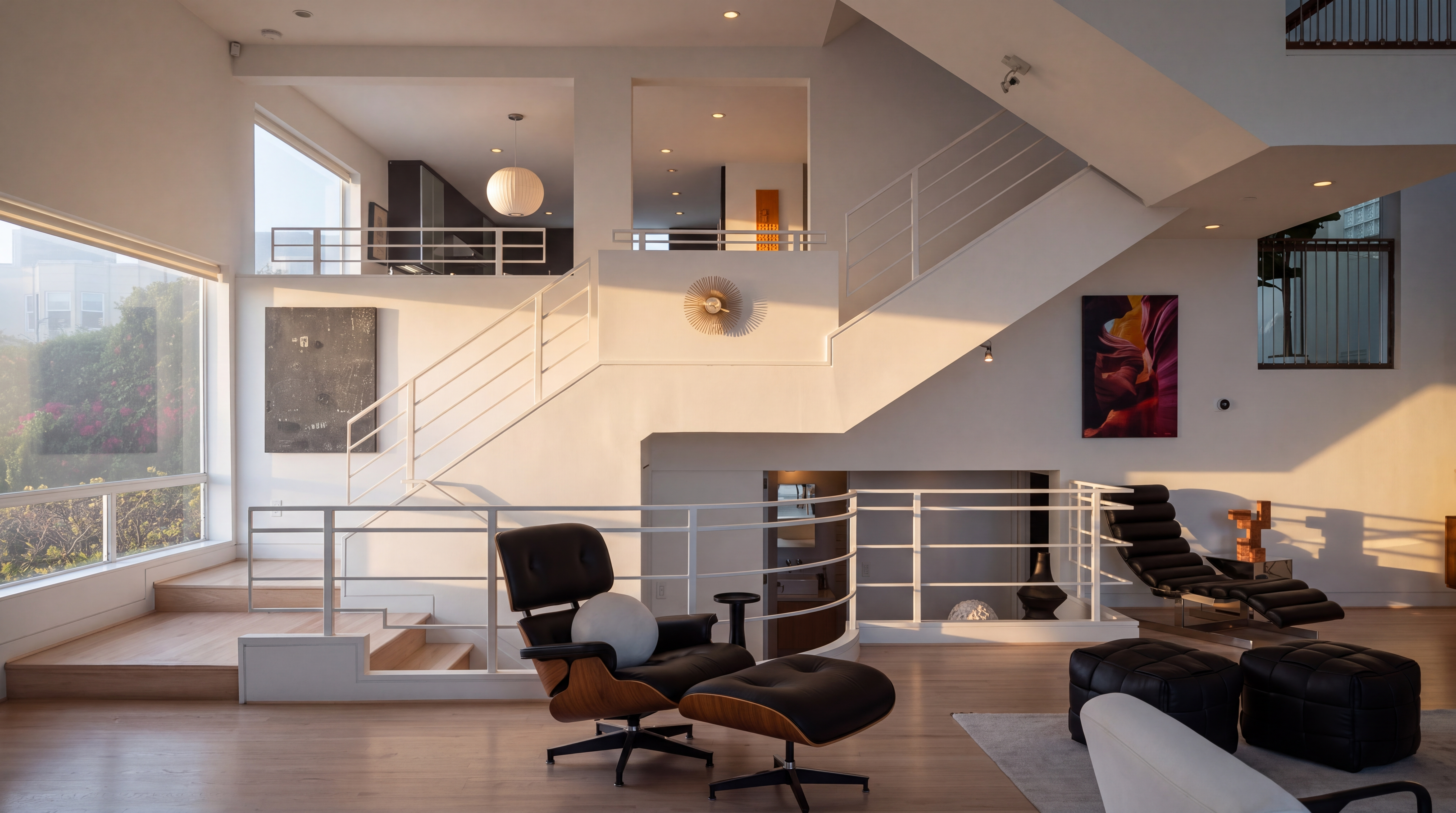 This expansive, multi-level living space features a modern, open-concept design with bright white walls and light wood flooring. A prominent white staircase with minimalist railings serves as a central architectural feature, connecting the main living area to an upper mezzanine level. The room is furnished with iconic mid-century modern lounge chairs, black leather ottomans, and bold abstract artwork, creating a sophisticated and airy atmosphere.