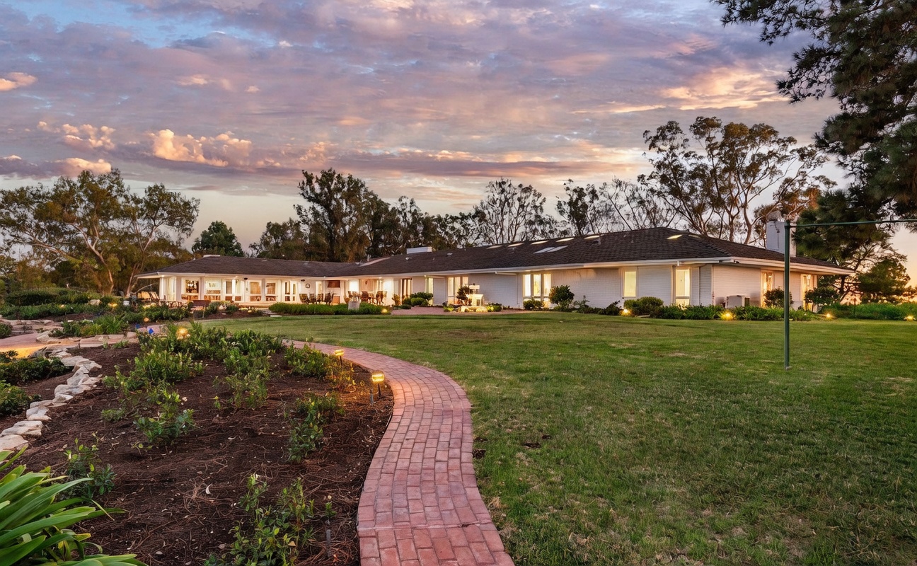This expansive single-story residence is captured from a wide-angle perspective, showcasing a sprawling backyard with a manicured lawn and a curved brick pathway leading toward the home. The house features a long, low-profile design with white exterior walls, a dark shingled roof, and numerous large windows that offer a clear view of the interior spaces. The scene is set during a vibrant sunset, with soft golden light illuminating the landscape and casting long shadows across the grass.