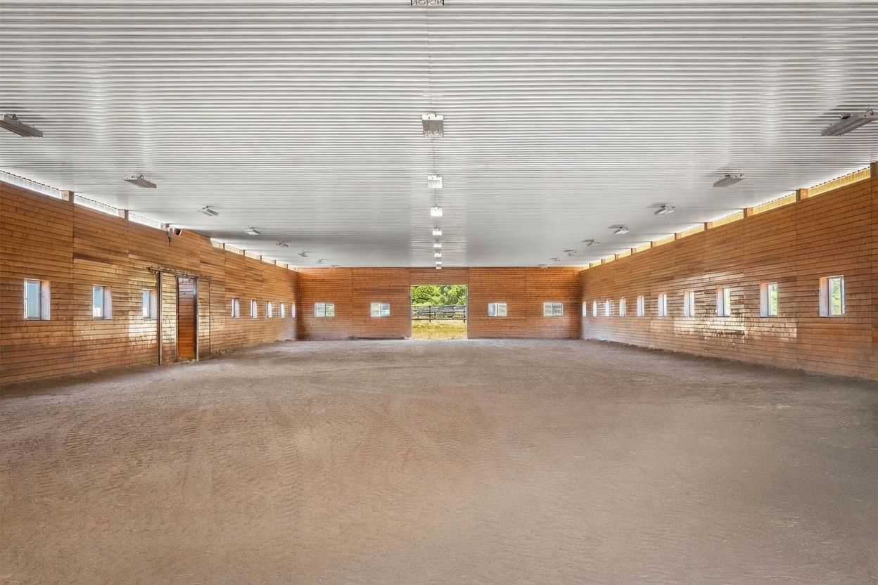 This image shows the interior of a large, open-concept indoor riding arena or multi-purpose barn structure. The space features high, corrugated metal ceilings with numerous overhead light fixtures and walls lined with horizontal wood planks. A dirt floor spans the entire area, leading to a central doorway that opens to an outdoor view, creating a sense of vast, functional space.