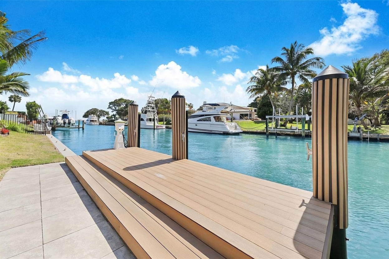 This image captures a serene waterfront perspective from a modern, light-toned composite deck overlooking a calm canal. Several yachts are moored in the distance, framed by lush tropical landscaping and a bright blue sky with scattered clouds. The scene emphasizes a luxurious, relaxed lifestyle with direct water access and a clear, sunny atmosphere.