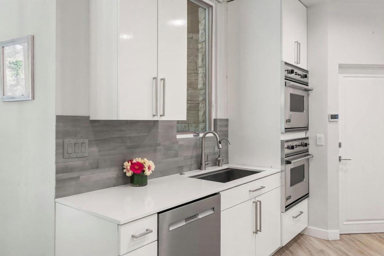 This modern kitchen features crisp white cabinetry paired with a sleek, light-colored countertop and a gray horizontal-tile backsplash. The space is equipped with stainless steel appliances, including a built-in double oven and a dishwasher, alongside a contemporary faucet and sink. A small window above the sink allows natural light to enter, creating a bright and functional culinary workspace.