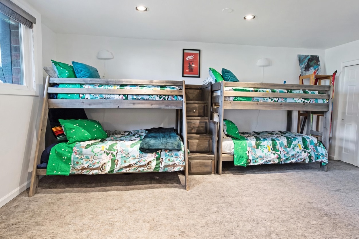 This bedroom features a custom-built, rustic-style wooden bunk bed configuration with an integrated staircase for access to the upper bunks. The room is decorated with playful, patterned bedding in shades of green and blue, complemented by neutral walls and carpeted flooring. A framed poster hangs on the wall between the two bunk units, adding a touch of character to this functional and space-efficient sleeping area.