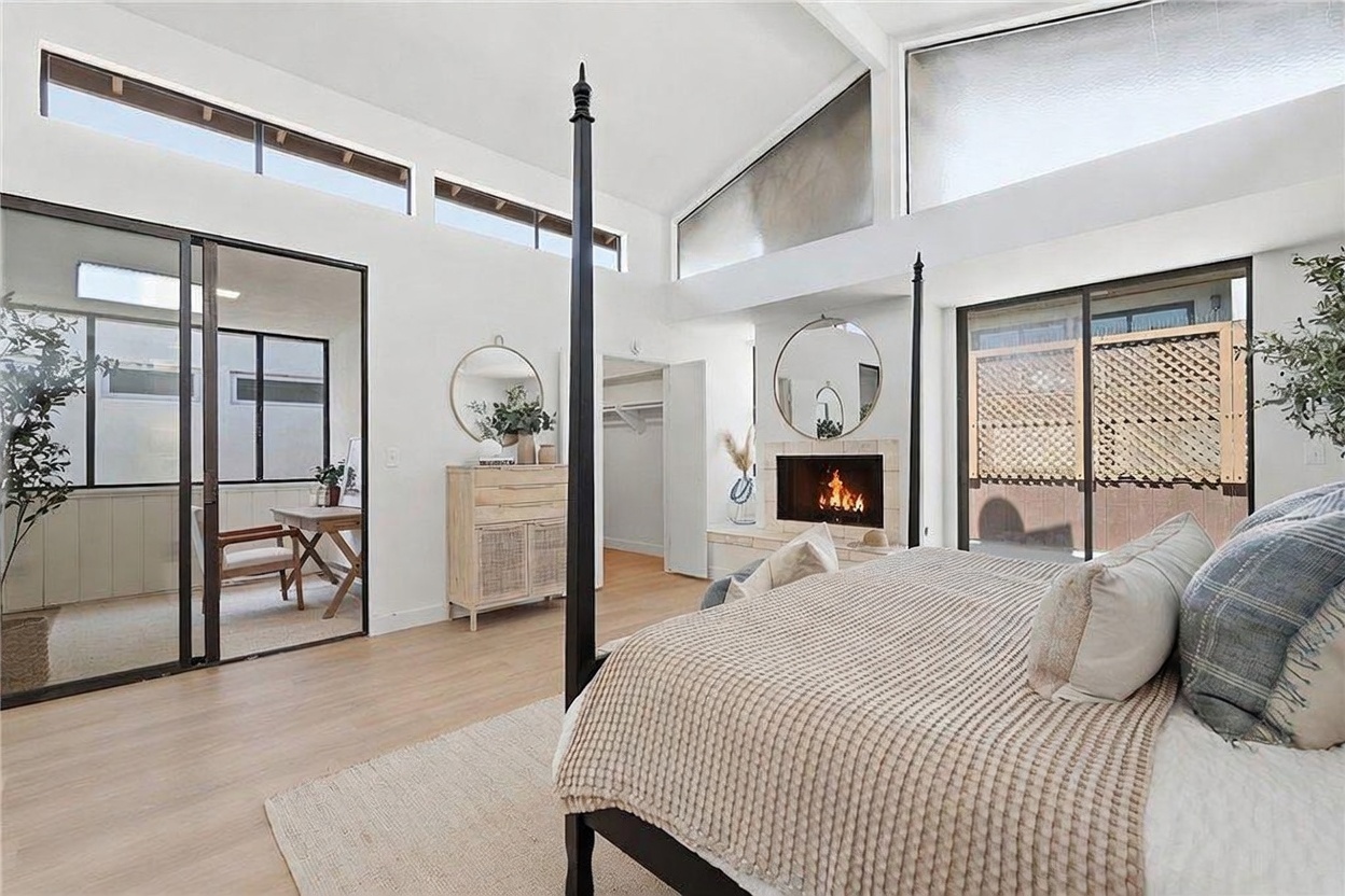 This spacious primary bedroom features a vaulted ceiling with exposed beams and large windows that allow for abundant natural light. The room is anchored by a four-poster bed and includes a cozy fireplace with a stone-clad chimney breast, alongside a light wood dresser and a mirror. The neutral color palette and light wood flooring create an airy, serene atmosphere, while sliding glass doors provide access to an outdoor space.