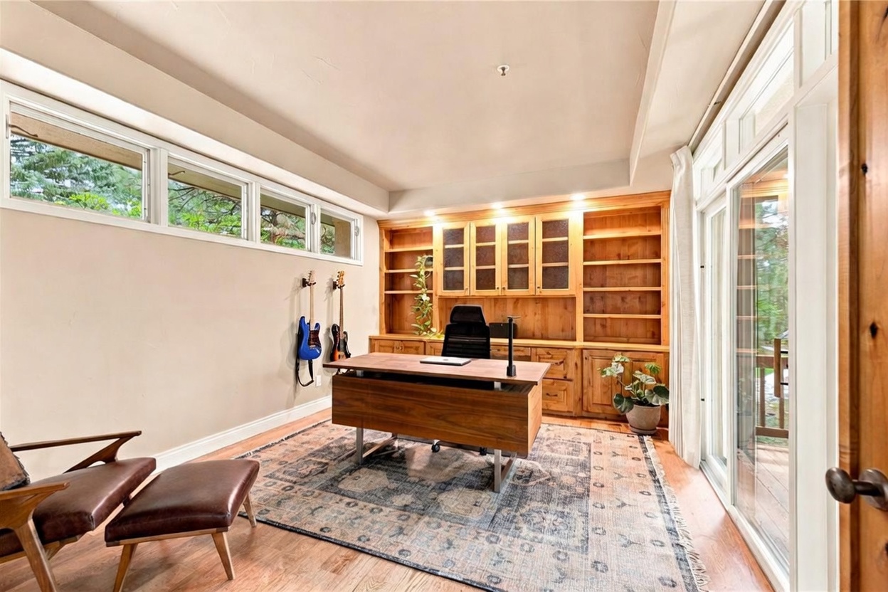 This home office features a large wooden desk positioned in the center of the room on a patterned area rug, facing a wall of built-in wooden cabinetry and shelving. To the left, two electric guitars are mounted on the wall beneath a row of horizontal windows, while a leather lounge chair and ottoman sit in the foreground. The room is bright and airy, with a sliding glass door on the right providing access to an outdoor deck and views of the surrounding trees.