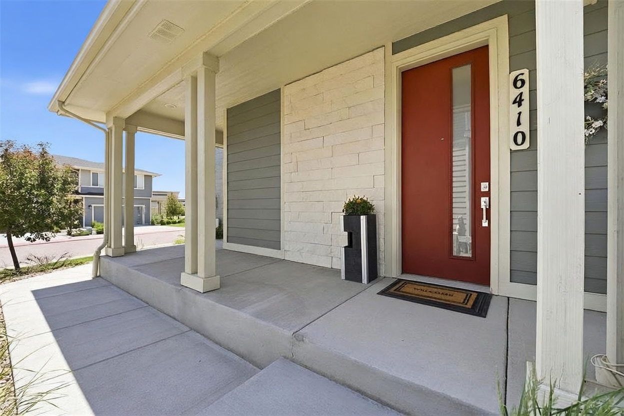 This inviting front entryway features a vibrant red door with a vertical glass panel, set against a backdrop of light-colored stacked stone and gray horizontal siding. A covered porch with white columns provides a sheltered transition, complemented by a welcome mat and a modern, tall planter. The perspective is a ground-level shot looking toward the entrance, highlighting the clean, contemporary curb appeal of the home.