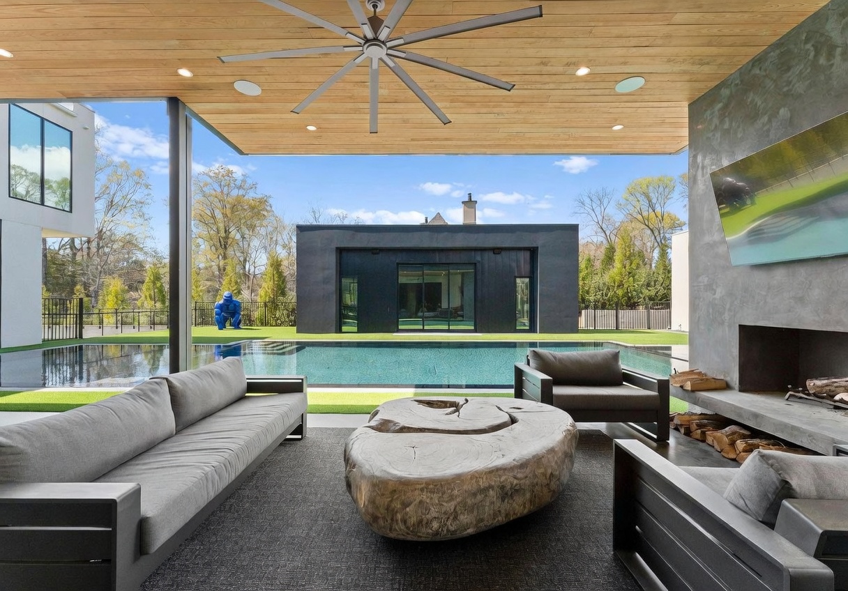 This luxurious covered outdoor living space features a modern aesthetic with a wood-paneled ceiling, a large ceiling fan, and a concrete fireplace with a mounted television. The space is furnished with contemporary grey seating and a unique, organic-shaped wooden coffee table, all set on a dark area rug. Beyond the patio, a pristine swimming pool reflects the clear blue sky, leading the eye toward a sleek, black-clad structure in the distance.