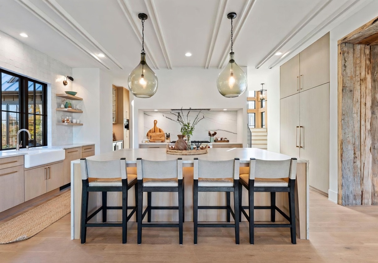 This bright, modern kitchen features a large central island with seating for four, topped with a light-toned surface and illuminated by two prominent glass pendant lights. The space showcases light wood cabinetry, a farmhouse-style sink, and a clean, minimalist aesthetic with a stone backsplash behind the range. The perspective is centered, highlighting the open-concept layout and the sophisticated architectural ceiling detail.