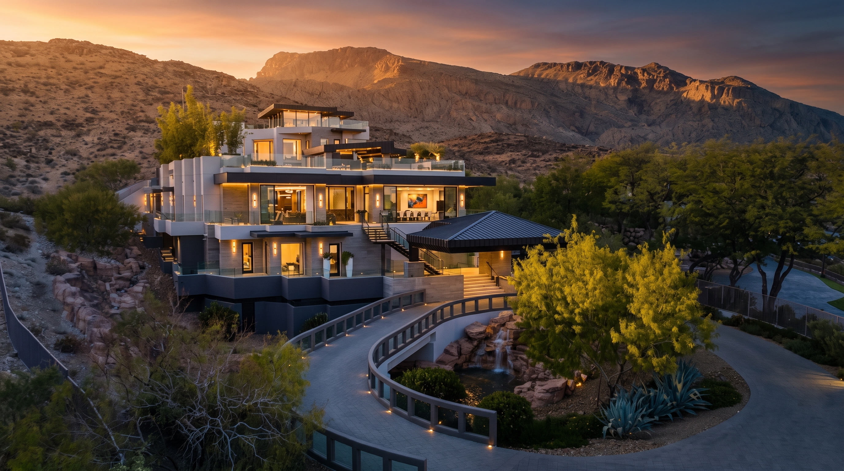 This multi-level contemporary luxury estate is nestled against a rugged mountain backdrop, showcasing a sophisticated architectural design with clean lines and expansive glass elements. The property features a sweeping, elevated driveway that leads to a covered entryway, complemented by a landscaped courtyard with a tiered water feature and mature trees. The structure utilizes a mix of stone and smooth-finish materials, creating a dramatic and imposing presence that integrates seamlessly with the desert landscape.