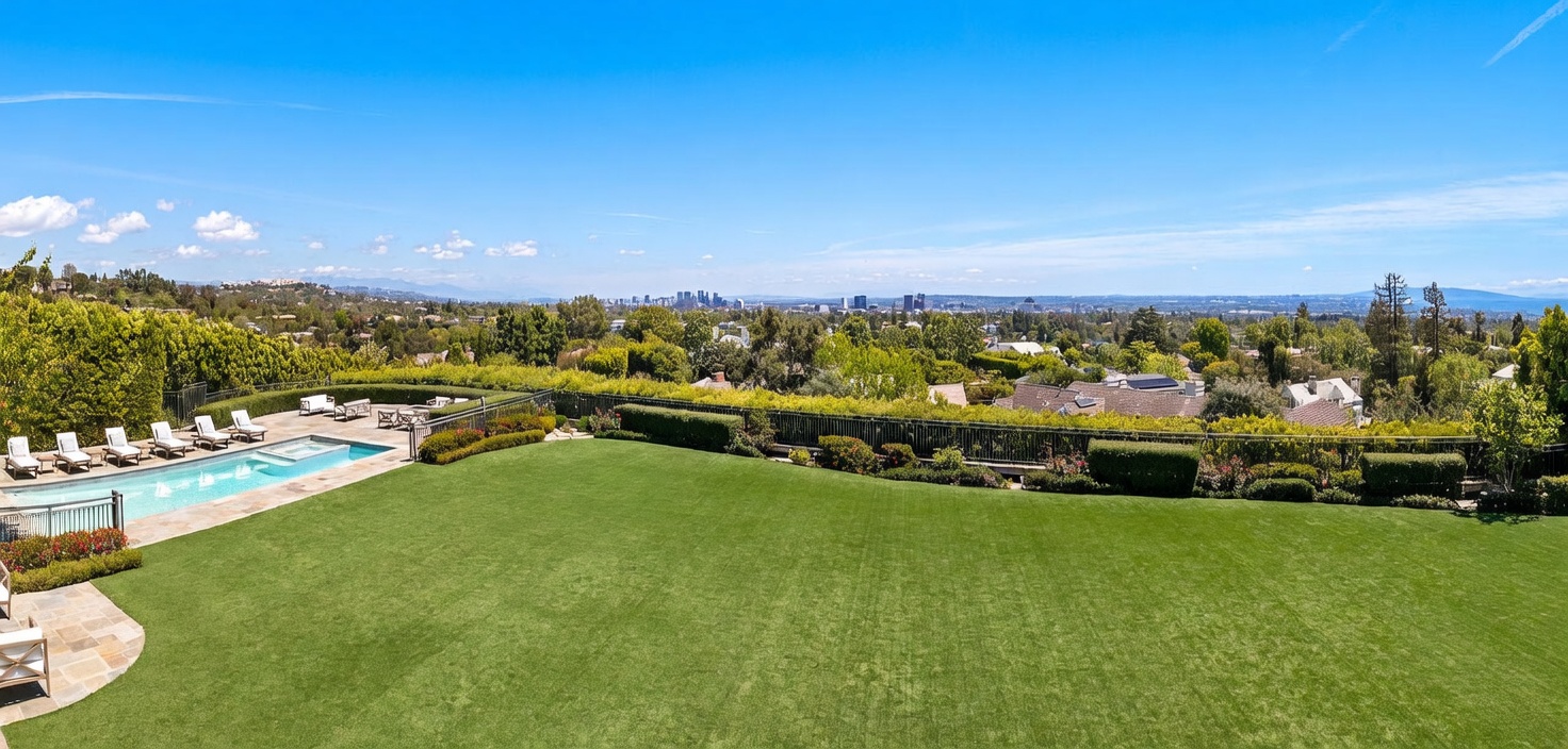 This expansive backyard features a meticulously manicured lawn that serves as the centerpiece, bordered by lush green hedges and a metal fence. To the left, a swimming pool with an integrated spa is surrounded by a stone patio furnished with lounge chairs, offering a perfect space for outdoor relaxation. The elevated perspective provides a sweeping, panoramic view of the surrounding neighborhood and the distant city skyline under a clear blue sky.