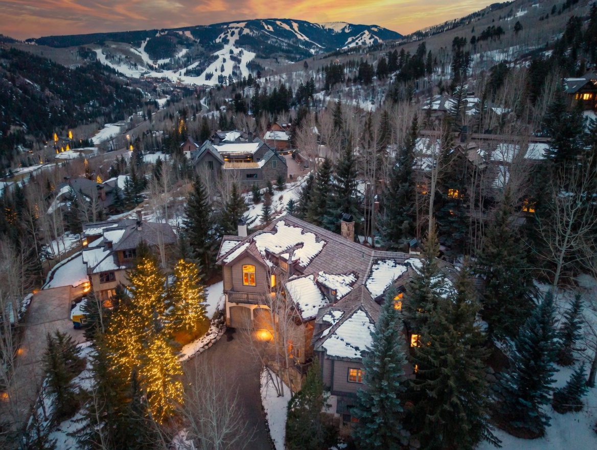 This high-angle aerial shot captures a luxurious mountain residence nestled within a dense, snow-dusted forest at dusk. The home features a multi-gabled roofline with patches of snow, warm interior lighting glowing through numerous windows, and a paved driveway illuminated by festive string lights on nearby trees. In the background, a majestic, snow-covered ski mountain rises against a vibrant sunset sky, creating a serene and upscale alpine atmosphere.