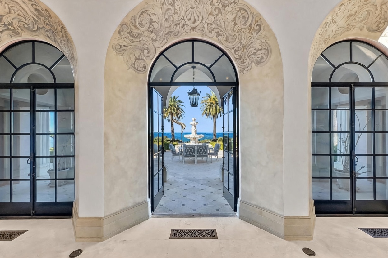 This elegant entryway features a grand arched doorway with black-framed glass doors that open to a scenic patio and ocean view. The interior space is framed by ornate, carved stone detailing above the arch, while the exterior patio showcases a classic fountain and outdoor dining set. The perspective looks directly through the threshold, creating a seamless transition from the refined indoor architecture to the expansive coastal landscape.