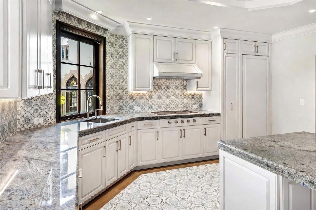 This elegant kitchen features white cabinetry paired with patterned tile backsplashes and dark stone countertops. The space includes a built-in refrigerator, a cooktop with a range hood, and a central island, all illuminated by recessed lighting. A window above the sink provides a view of the exterior, contributing to a bright and sophisticated atmosphere.