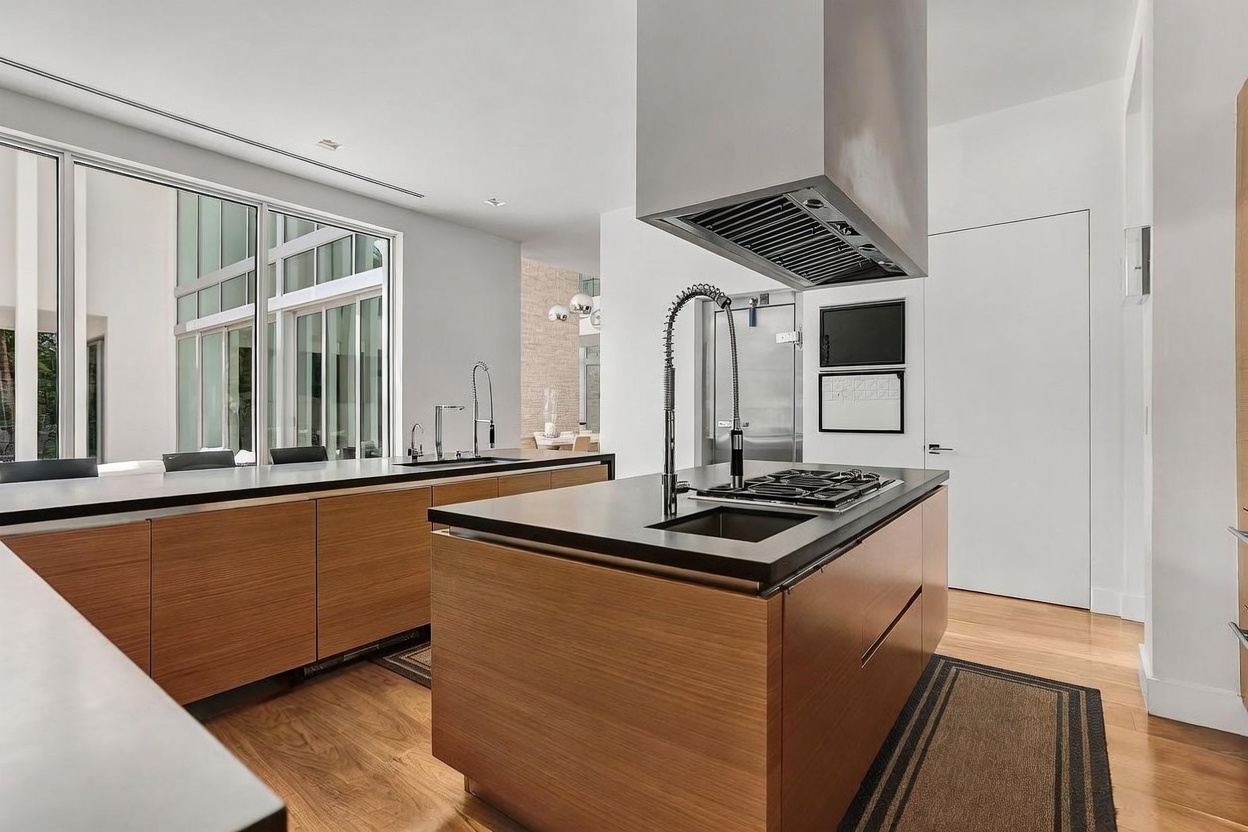 This modern kitchen features a large central island with a dark countertop, a built-in cooktop, and a prominent stainless steel range hood suspended from the ceiling. The cabinetry is finished in a warm-toned wood, complemented by light wood flooring and expansive windows that provide ample natural light. The space is designed with a clean, minimalist aesthetic, offering a clear view into an adjacent living area.