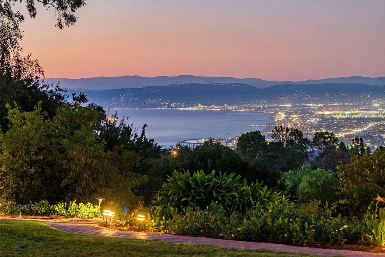 This image captures a breathtaking twilight view from a landscaped garden overlooking a coastal city and bay. In the foreground, a paved stone path winds through lush greenery and manicured shrubs, illuminated by warm, low-level garden lighting. The background features a panoramic vista of a glowing cityscape reflecting over the water, framed by distant mountain silhouettes under a soft, gradient sunset sky.