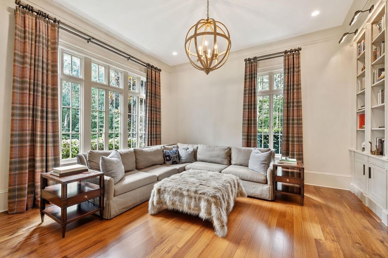 This elegant living room features a large, neutral-toned sectional sofa arranged around a plush, textured ottoman on a patterned area rug. The space is anchored by a prominent spherical chandelier and flanked by tall windows dressed in plaid curtains, which allow ample natural light to fill the room. To the right, a built-in shelving unit provides ample storage and display space, contributing to the sophisticated and comfortable atmosphere of the home.