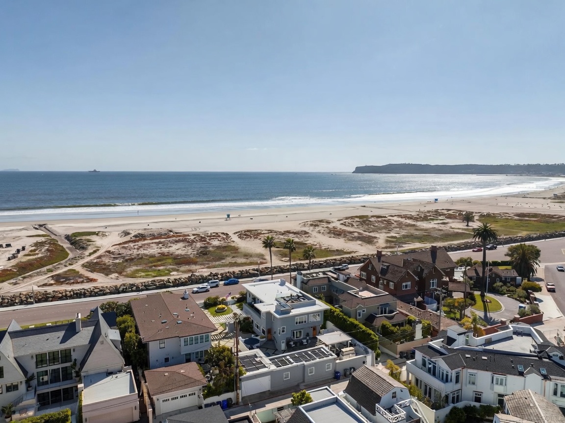 This high-angle aerial view captures a coastal neighborhood situated directly adjacent to a wide, sandy beach and the Pacific Ocean. The scene highlights a collection of multi-story residential homes with varied architectural styles, including flat-roofed modern structures and traditional pitched-roof houses. The expansive horizon, clear blue sky, and proximity to the shoreline emphasize the prime, desirable location of these properties.