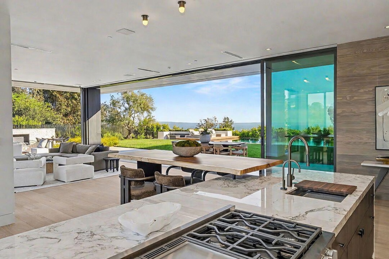 This open-concept kitchen features a large island with a stone countertop and a built-in gas cooktop, seamlessly transitioning into a dining area with a natural wood table. The space is defined by expansive, retractable glass walls that open completely to an outdoor patio, pool, and lush greenery, creating a fluid indoor-outdoor living experience. The interior design is modern and minimalist, characterized by clean lines, neutral tones, and recessed ceiling lighting.