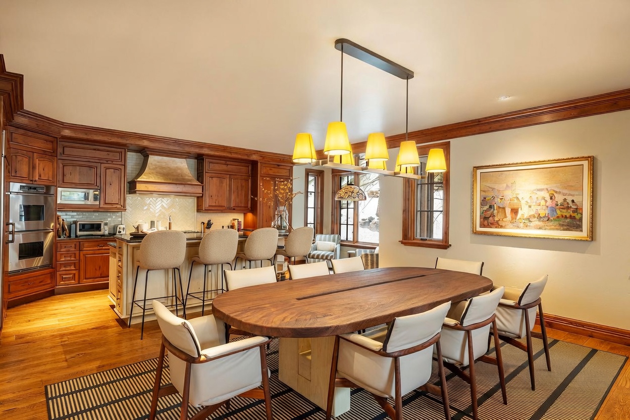 This open-concept space features a large, organic-shaped wooden dining table surrounded by modern chairs, positioned adjacent to a well-equipped kitchen. The kitchen showcases warm-toned cabinetry, a stone-tiled backsplash, and a breakfast bar with stools, creating a cohesive and inviting atmosphere. Large windows allow natural light to fill the room, which is accented by a multi-pendant light fixture and a framed piece of art on the wall.