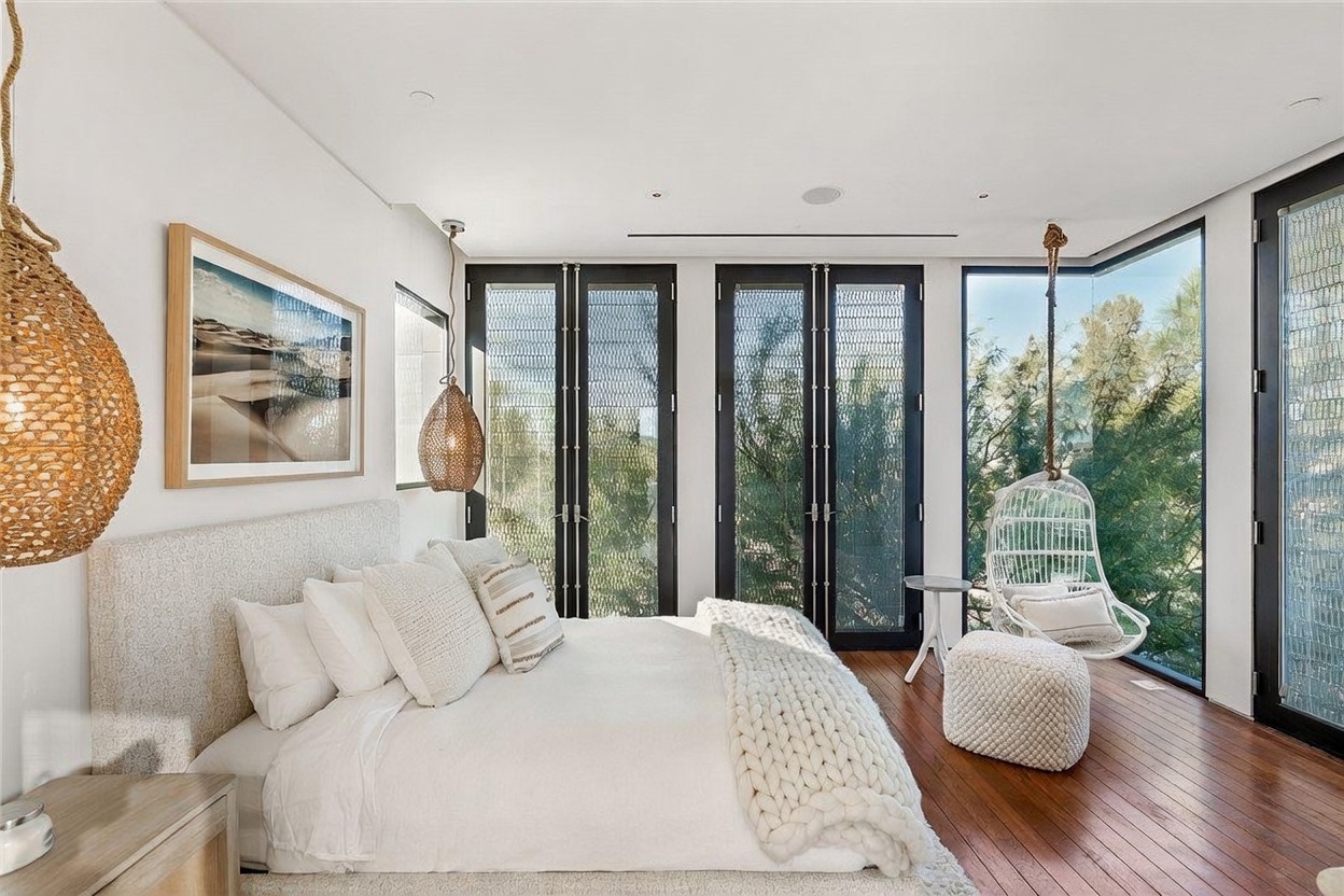 This bright and airy primary bedroom features a neutral color palette with a plush, textured bed as the central focal point. The room is accented by woven pendant lights and a hanging chair positioned near large windows that provide an expansive view of the surrounding greenery. The space is finished with warm wood flooring, creating a serene and sophisticated atmosphere.