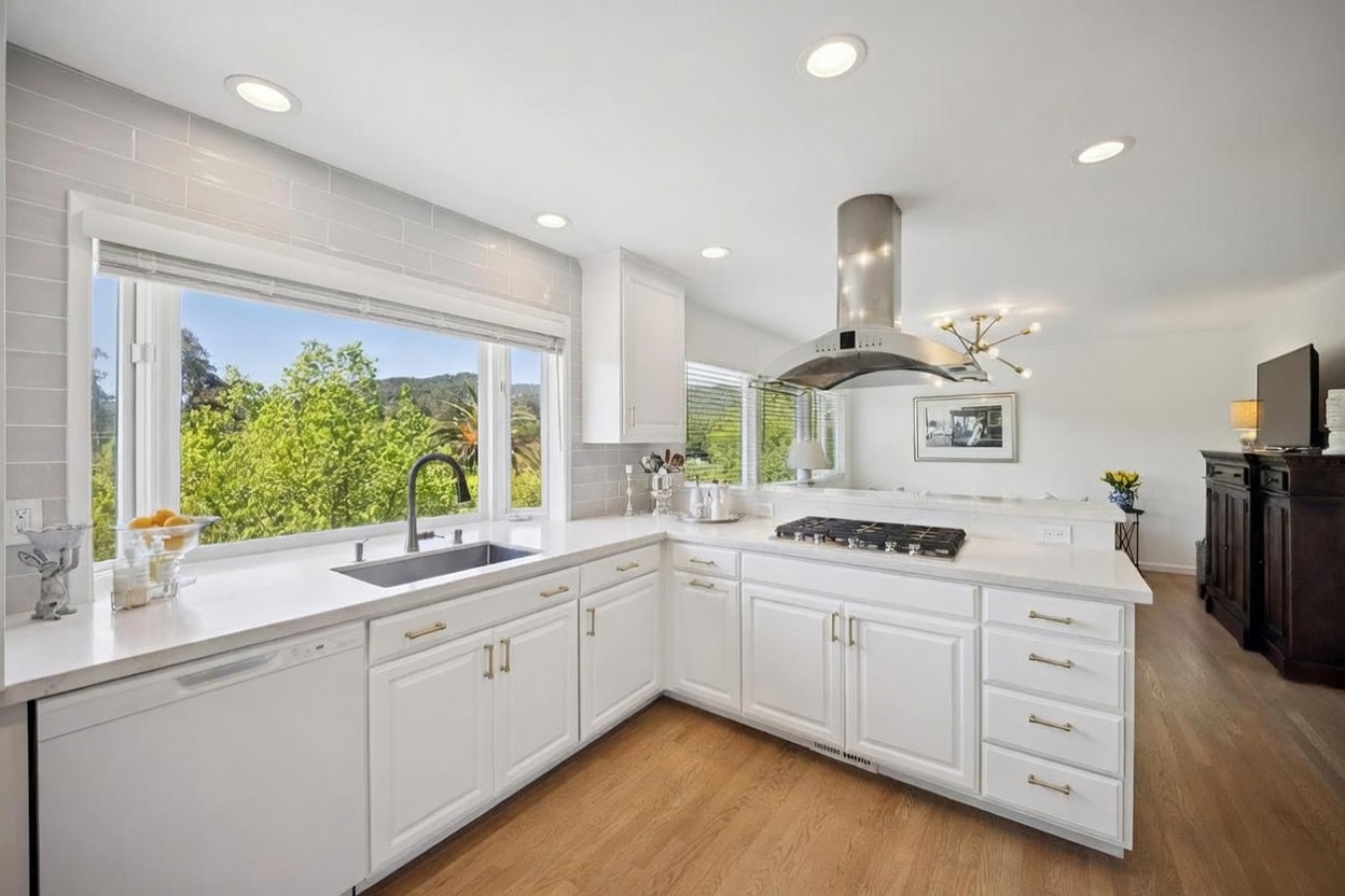 This bright and airy kitchen features crisp white cabinetry, light-toned countertops, and a large window above the sink that provides a scenic view of the surrounding greenery. A stainless steel range hood hangs above the cooktop, which is set into an L-shaped counter configuration with light wood flooring throughout. The space is illuminated by recessed lighting and a modern chandelier in the adjacent dining area, creating a clean and inviting atmosphere.