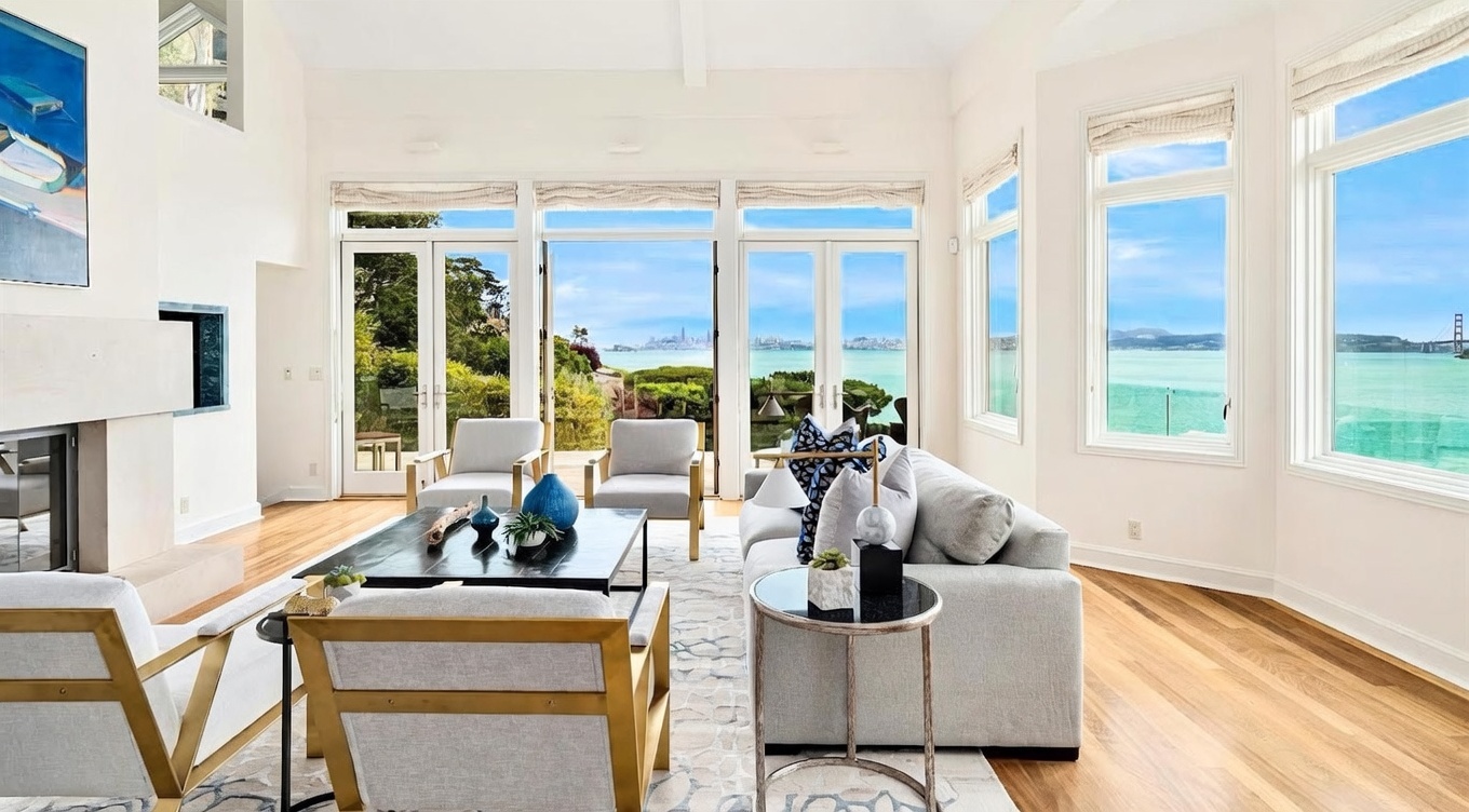 This bright and airy living room features a neutral color palette with light-toned wood flooring and a large, patterned area rug. The space is anchored by a comfortable sofa and several armchairs, centered around a dark coffee table, with a tall fireplace surround on the left. Large glass doors and windows provide an expansive, unobstructed view of the bay and distant city skyline, creating a seamless connection between the indoor living space and the scenic outdoors.