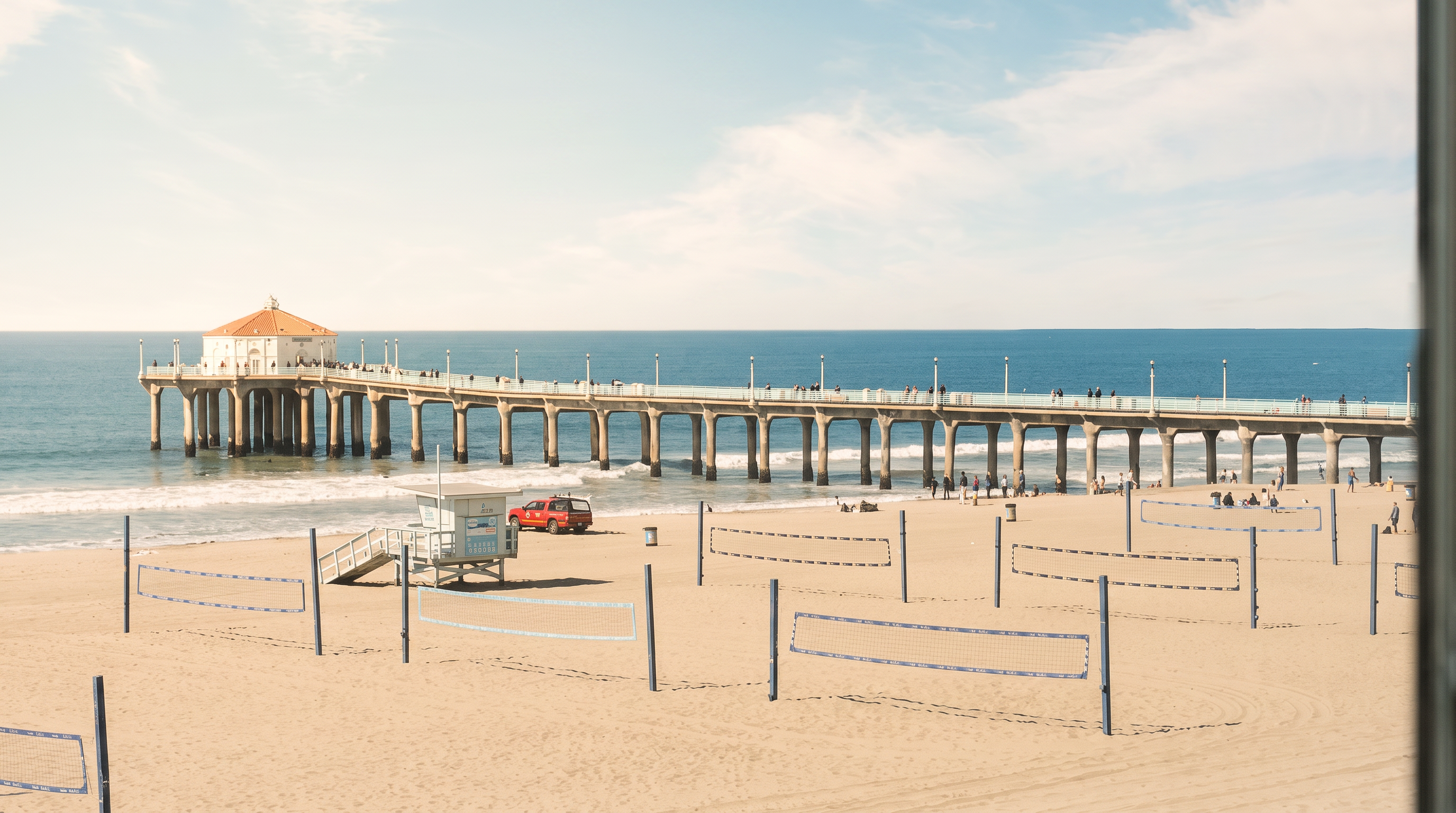 This view is captured from an elevated vantage point, likely a balcony or window, looking out over a sandy beach and the iconic Manhattan Beach Pier. The scene features multiple volleyball nets set up on the sand, with the vast blue ocean stretching toward the horizon under a clear, bright sky. The perspective provides a serene and expansive coastal atmosphere, highlighting the property's prime location near a popular recreational landmark.