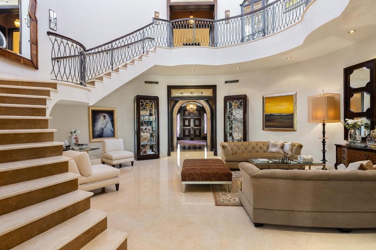 This grand foyer features a sweeping, curved staircase with ornate iron railings that serves as the focal point of the space. The open-concept area flows into a sophisticated living room furnished with plush, neutral-toned seating and elegant decor. High ceilings, polished marble flooring, and a symmetrical layout create an atmosphere of luxury and refined architectural design.
