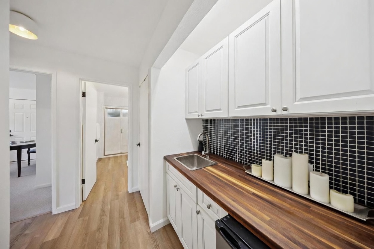 This interior view showcases a compact wet bar or kitchenette area featuring white cabinetry paired with a warm-toned wood countertop and a striking black tiled backsplash. A stainless steel sink is integrated into the counter, which is accented by a collection of pillar candles on a decorative tray. The perspective looks down a hallway toward an adjacent room, highlighting the clean, modern aesthetic and the transition between living spaces.