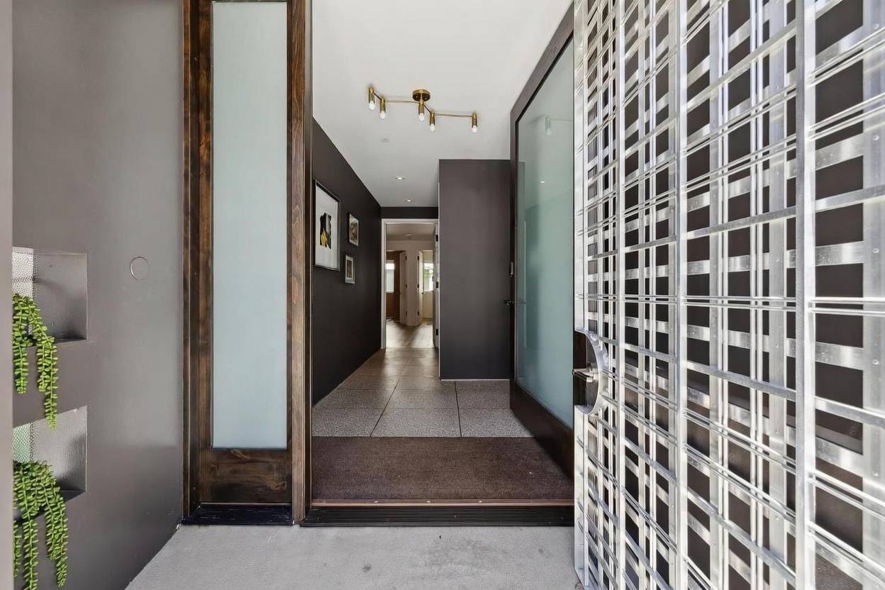 This entryway features a striking metal security gate with a geometric grid pattern that opens into a modern foyer. The interior hallway is defined by dark walls, a light-colored tiled floor, and a minimalist gold-toned light fixture. A frosted glass door panel provides privacy while allowing natural light to filter into the home, creating a sophisticated and welcoming transition from the exterior.