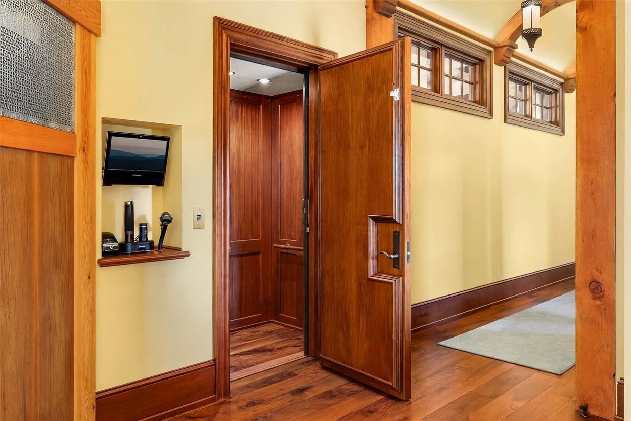 This interior view captures a hallway featuring rich wood trim and flooring, leading to a private elevator with wood-paneled walls. To the left, a recessed niche houses a small monitor and electronic equipment, while the right side features a long wall with windows that allow natural light to filter into the space. The warm wood tones and classic architectural details create a sophisticated and inviting atmosphere.