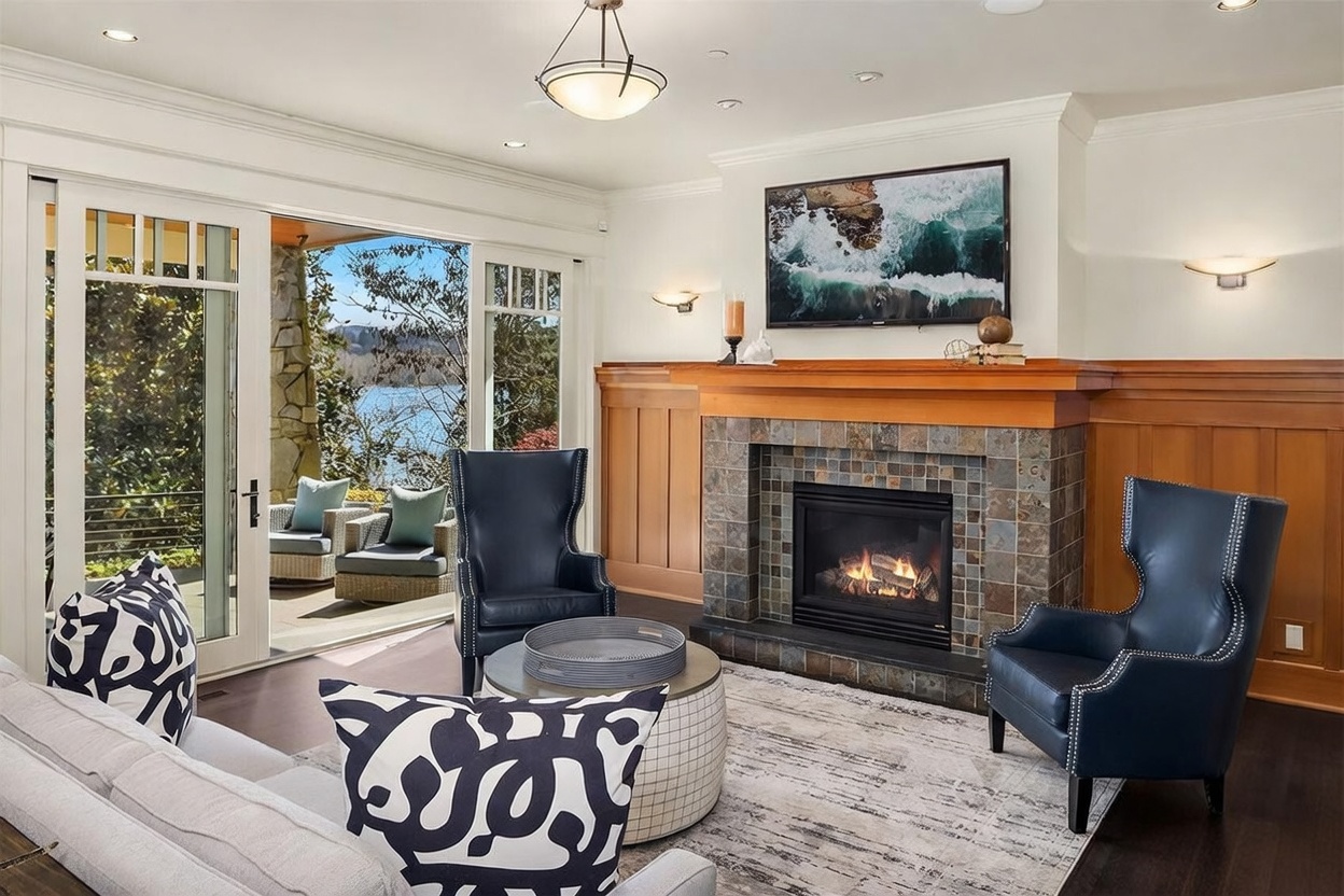 This elegant living room features a cozy fireplace with a stone surround and a wooden mantel, serving as the focal point of the space. The room is furnished with two dark blue leather wingback chairs and a plush sofa, all arranged around a circular ottoman on a textured area rug. Large sliding glass doors open to a scenic outdoor patio, seamlessly blending indoor comfort with beautiful waterfront views.