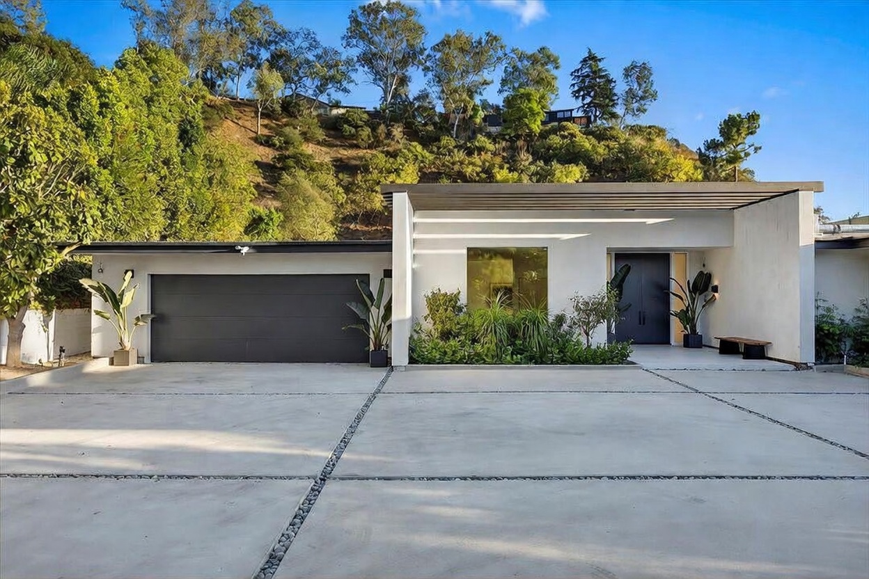 This modern, single-story residence features a clean, minimalist aesthetic with a prominent dark garage door and a recessed entryway. The facade is characterized by crisp white walls, a flat roofline, and a large, square window that provides a focal point near the front door. A wide concrete driveway leads up to the house, which is set against a lush, hillside backdrop, creating a serene and private atmosphere.