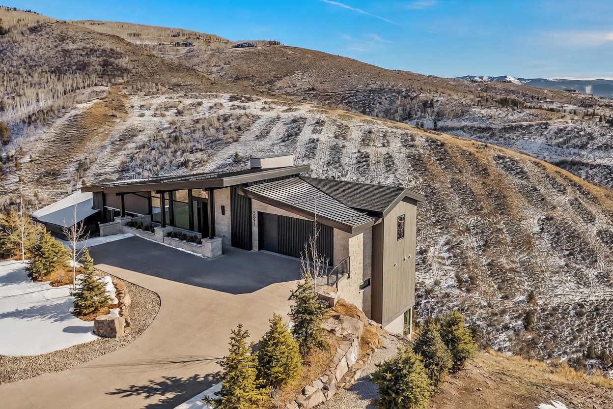 This image showcases the front exterior of a modern home nestled on a hillside. The architectural design features a combination of stone and wood siding, with a sleek, angled roofline. A concrete driveway leads up to the garage, and the surrounding landscape includes mature trees and natural terrain, creating a sense of privacy and integration with the environment.