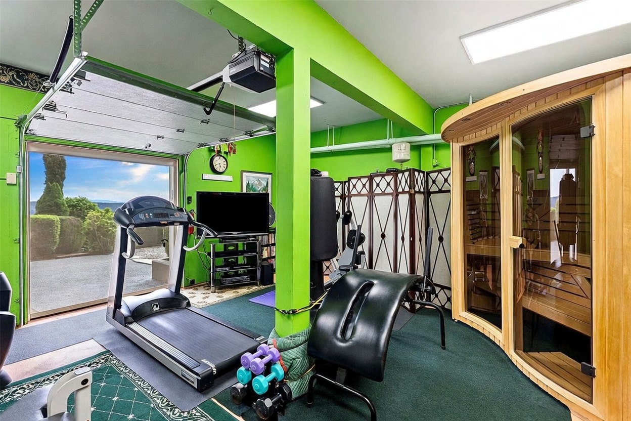 This vibrant home gym is situated within a converted garage space, featuring bright green walls and a green carpeted floor. The room is equipped with a treadmill, a weight bench, a sauna, and various free weights, creating a functional workout area. A large open garage door reveals a scenic outdoor view, blending the indoor fitness space with the natural surroundings.