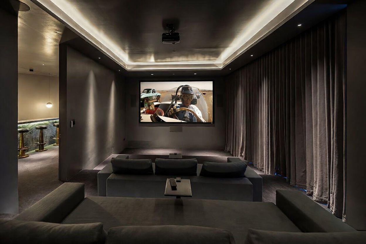 This image showcases a sophisticated home theater room featuring tiered seating with plush, dark-toned sofas and a large projection screen displaying a classic racing film. The room is designed with recessed cove lighting in the ceiling, dark wall treatments, and heavy curtains along the side wall to enhance the cinematic experience. To the left, a glimpse of an adjacent bar area with gold-accented stools adds a touch of luxury to the entertainment space.