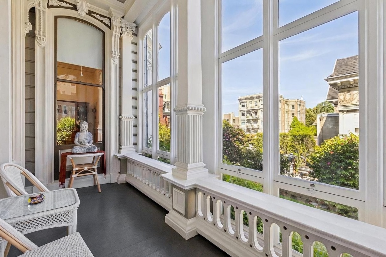 This bright and airy sunroom features classic architectural details, including ornate white columns and a decorative balustrade. Large windows provide an expansive view of the surrounding neighborhood, filling the space with natural light. The room is furnished with light-toned wicker seating and a decorative Buddha statue, creating a serene and peaceful atmosphere.