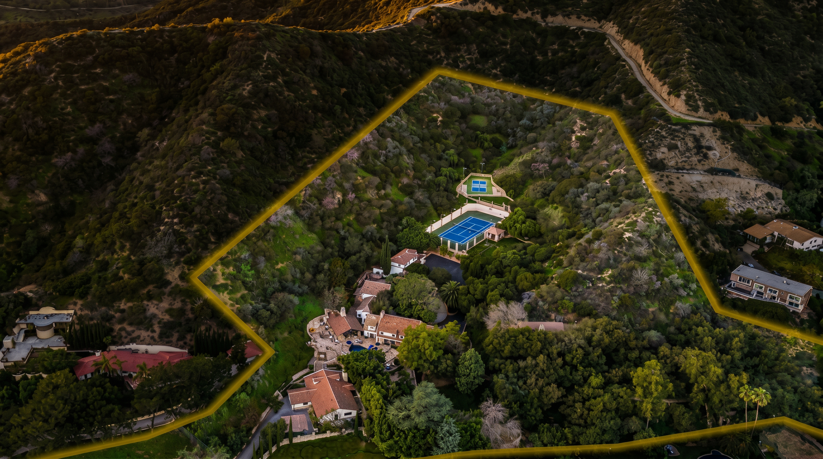 This high-angle aerial view captures a sprawling, secluded estate nestled within a lush, hilly landscape. The property features a large main residence with multiple structures, a private tennis court, and extensive wooded grounds enclosed by a highlighted boundary line. The perspective emphasizes the expansive nature of the lot and its integration into the natural terrain, offering a sense of privacy and luxury.