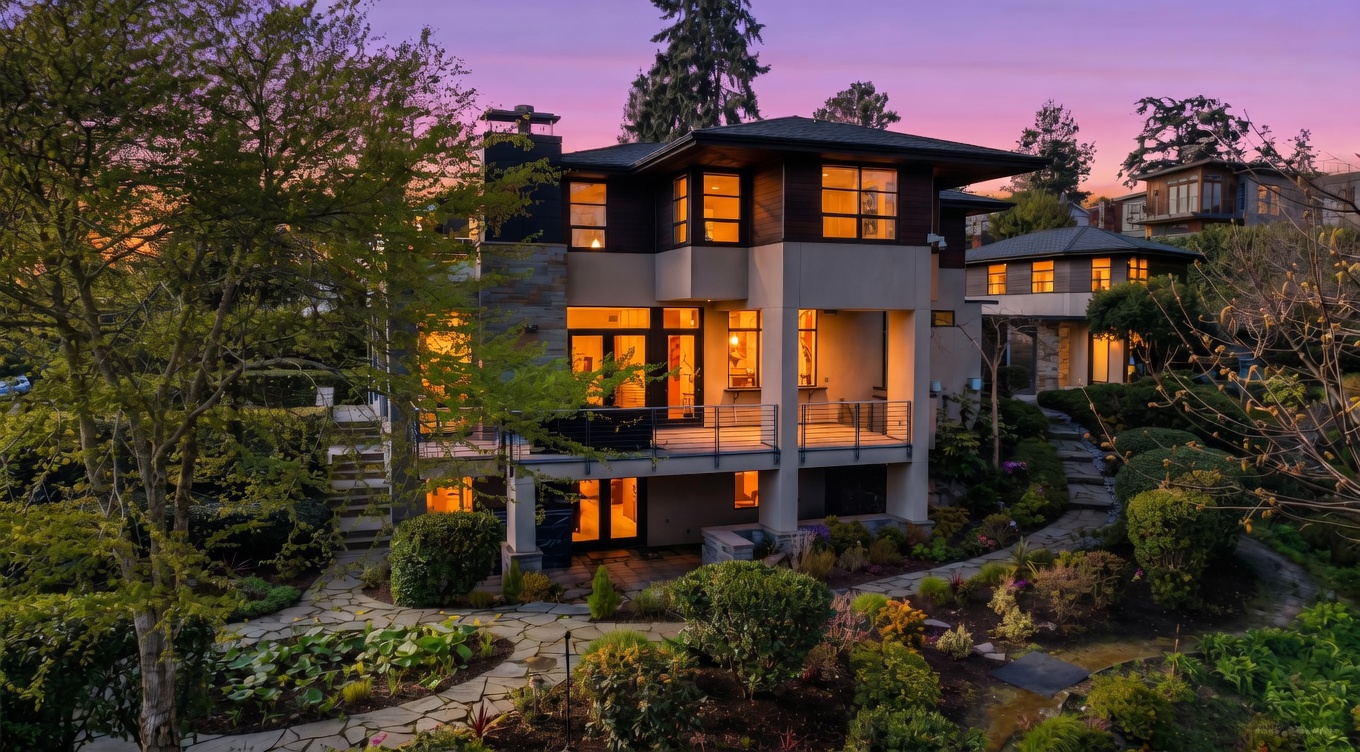This contemporary multi-level residence is captured at dusk, showcasing a sophisticated architectural design with clean lines and a mix of stone and smooth exterior finishes. The home features a spacious elevated deck overlooking a lush, landscaped garden with winding stone pathways and mature trees. The warm glow from the interior lighting creates an inviting atmosphere against the vibrant purple and orange hues of the twilight sky.