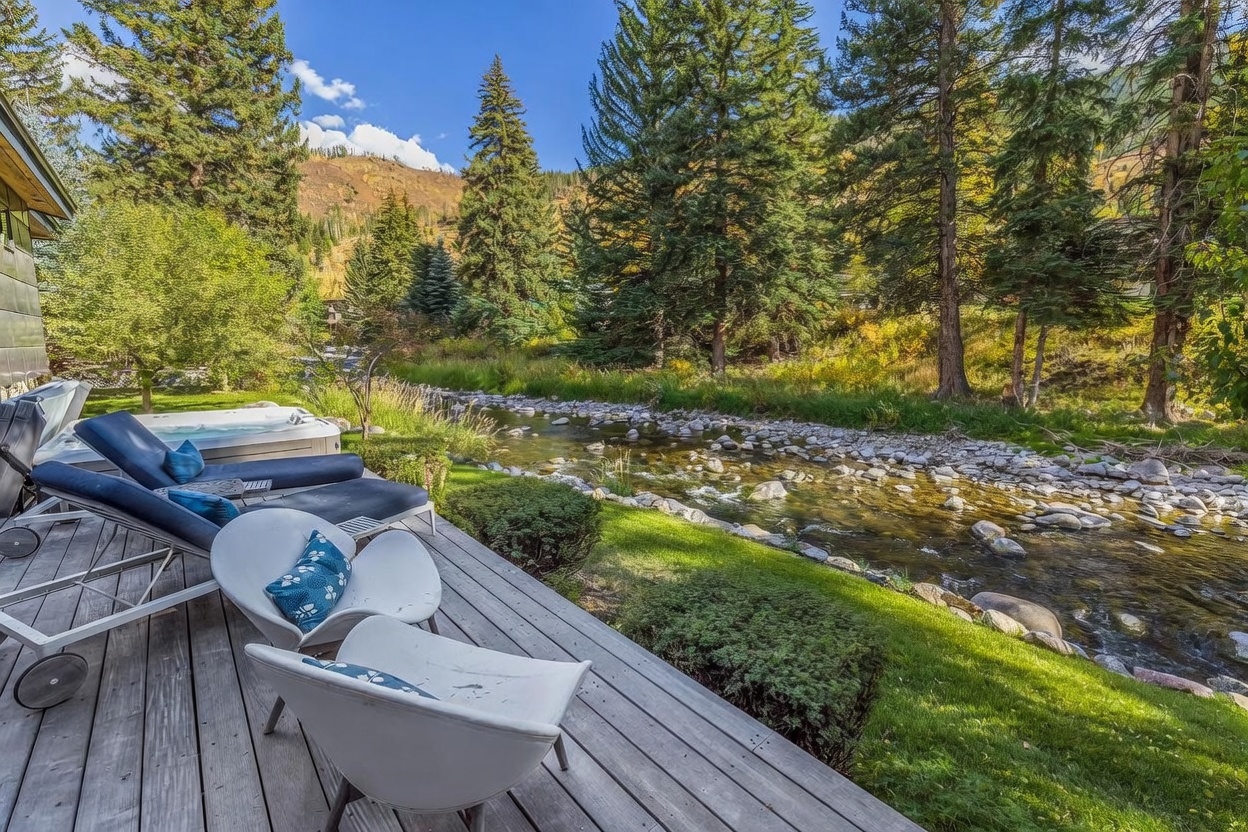 This serene outdoor deck offers a tranquil retreat overlooking a rushing mountain stream. The space is furnished with two modern lounge chairs and two white accent chairs, complemented by a private hot tub situated at the edge of the wooden decking. Tall evergreen trees and a lush, natural landscape frame the view, creating a peaceful and secluded atmosphere.