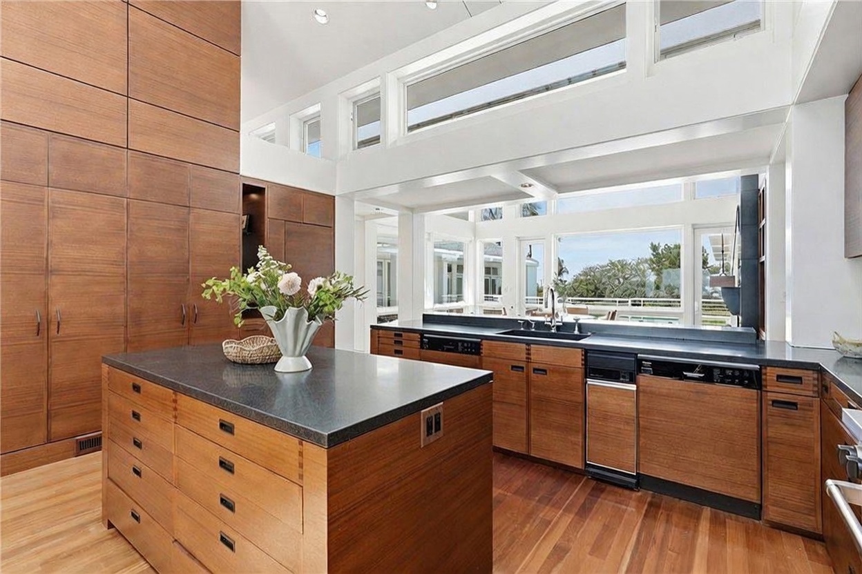 This modern kitchen features warm-toned wood cabinetry and a large central island topped with a dark stone countertop. The space is bright and airy, highlighted by a series of clerestory windows that allow natural light to flood the room. A view through the large windows above the sink reveals an outdoor deck area, creating a seamless connection between the indoor and outdoor living spaces.