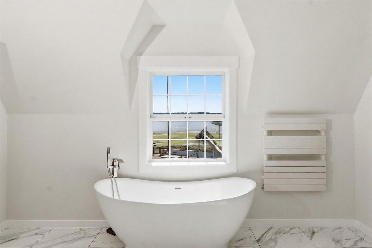 This bright and airy bathroom features a sleek, freestanding white soaking tub positioned directly beneath a window that offers a scenic view of the water. The space is finished with light-colored, patterned tile flooring and a wall-mounted towel warmer, creating a clean and minimalist aesthetic. The vaulted ceiling adds architectural interest, enhancing the sense of openness and tranquility in this spa-like retreat.