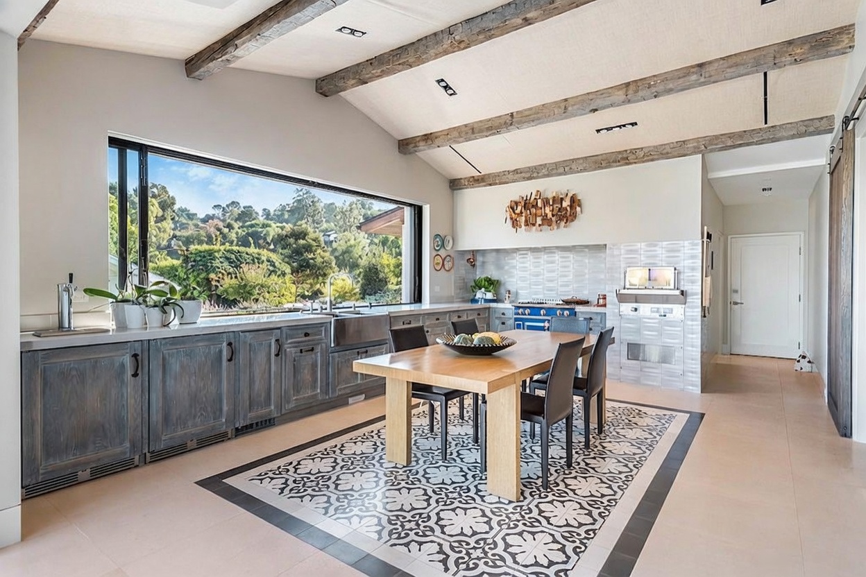 This spacious kitchen features a rustic-modern aesthetic with exposed wooden ceiling beams and a large, central dining table set on a decorative patterned tile floor. A long, expansive window provides a panoramic view of the lush outdoor landscape, while the cabinetry is finished in a weathered, textured gray tone. The space is bright and airy, blending natural elements with contemporary design to create a warm, inviting atmosphere.