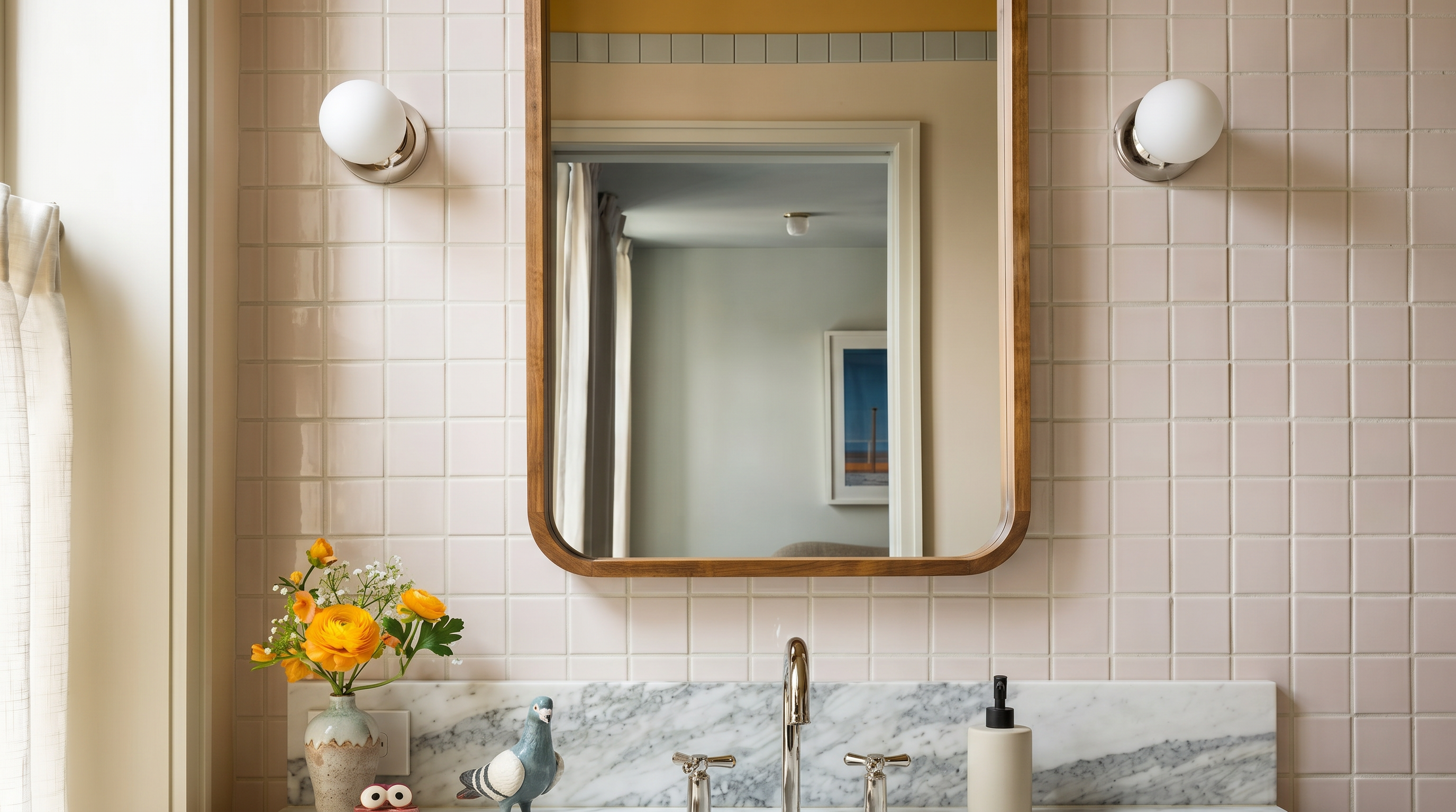 This elegant bathroom vanity features a rich wood cabinet topped with a white marble countertop and a classic undermount sink. The wall is adorned with light-colored square tiles, accented by a marble backsplash and a stylish, rounded-edge wooden mirror flanked by two globe-style wall sconces. A small vase of yellow flowers and a decorative bird figurine add a touch of personality to this well-appointed, warm, and inviting space.