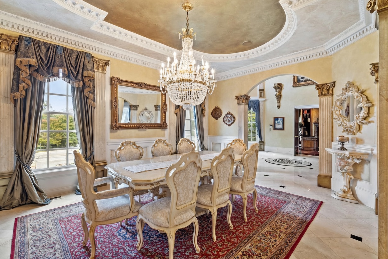This formal dining room exudes opulence with its ornate crystal chandelier, coffered ceiling, and classic architectural columns. A large, light-toned dining table is surrounded by six upholstered chairs, all set atop a rich, patterned area rug. The space is framed by tall windows with heavy drapery and features decorative wall mirrors and a console table, creating a sophisticated and traditional atmosphere.