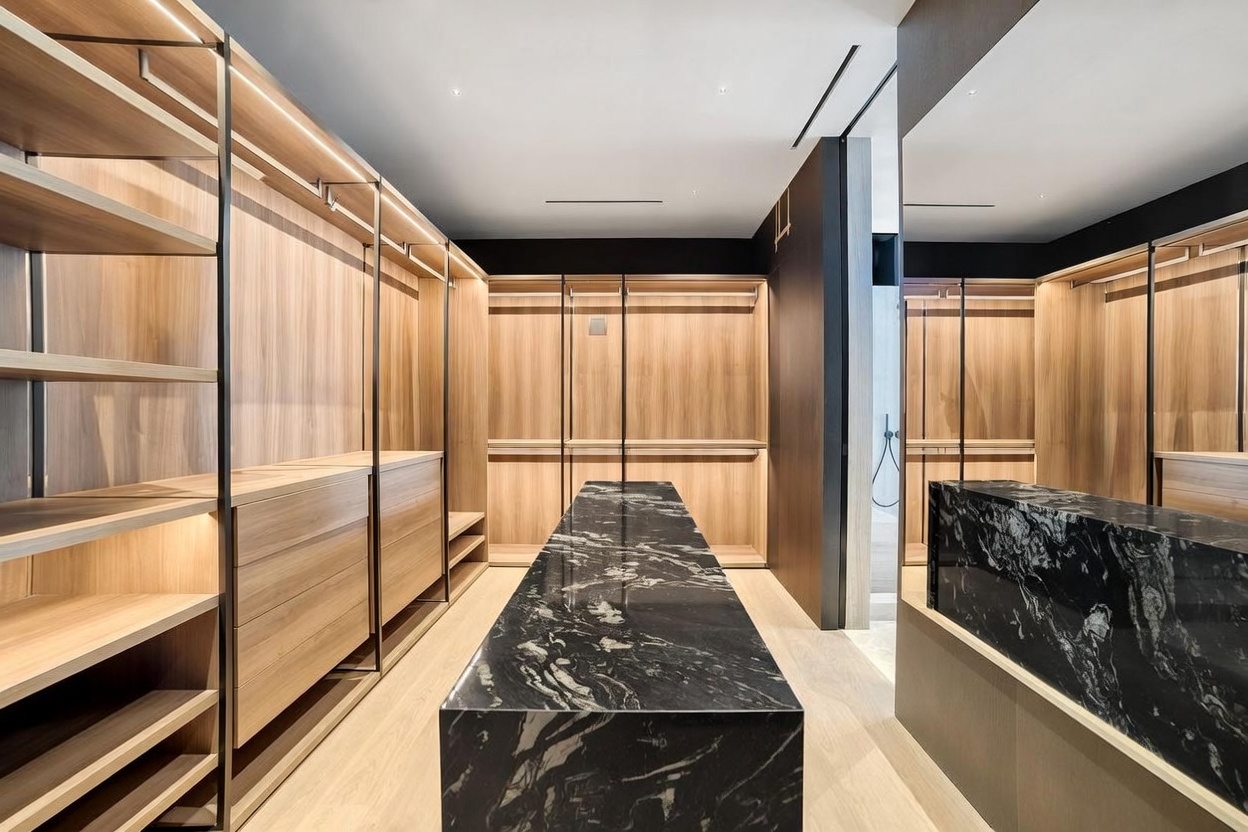This sophisticated walk-in closet features extensive light wood cabinetry with integrated hanging rods and shelving. A central island topped with a striking dark, veined stone provides a bold focal point, while a large mirror on the right wall enhances the sense of depth and space. The room is illuminated by recessed lighting, creating a clean, modern, and luxurious atmosphere.
