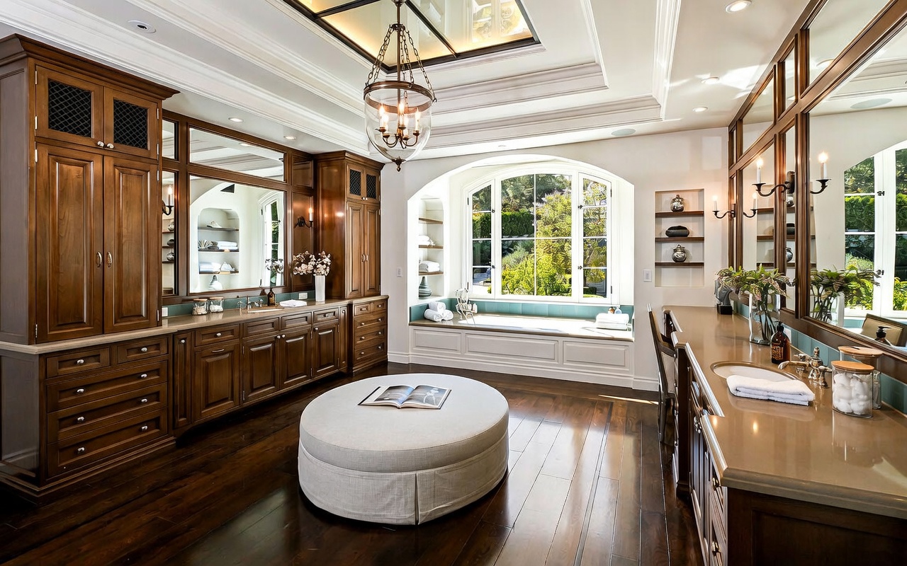 This luxurious primary bathroom features rich, dark wood cabinetry, dual vanities with stone countertops, and a central circular ottoman. A deep soaking tub is positioned beneath a large arched window that overlooks lush greenery, while a decorative chandelier hangs from the coffered ceiling. The space is finished with dark wood flooring and elegant wall sconces, creating a sophisticated and tranquil atmosphere.