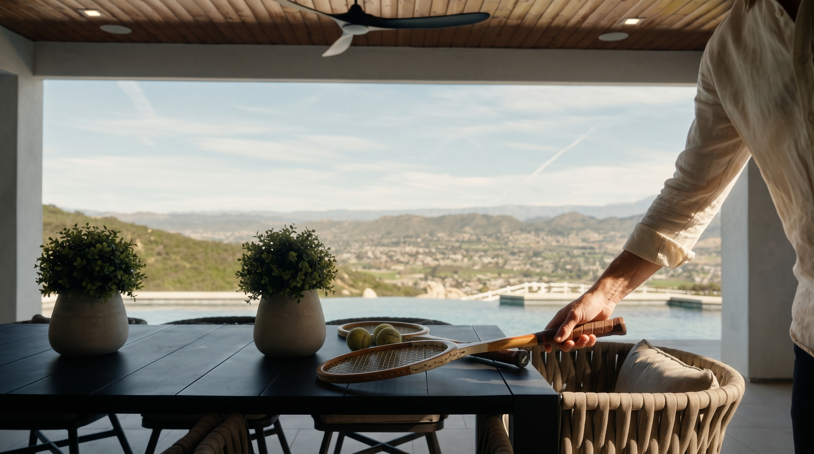 This covered outdoor patio features a contemporary dining setup with a dark wooden table and woven-style chairs, perfect for alfresco dining. The space opens up to an impressive infinity-edge pool that seamlessly blends with the panoramic valley and mountain views in the background. The area is highlighted by a high, natural wood ceiling with a modern fan, creating an inviting and luxurious indoor-outdoor living experience.