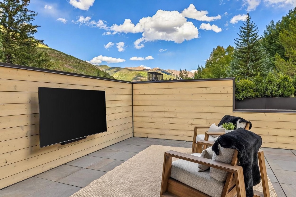 This modern outdoor terrace features light wood-paneled walls and dark tile flooring, creating a sleek, contemporary aesthetic. The space is furnished with two comfortable armchairs draped with dark faux-fur throws, positioned to face a wall-mounted television. The open-air setting offers a stunning, unobstructed view of the surrounding mountain range under a bright, blue sky.